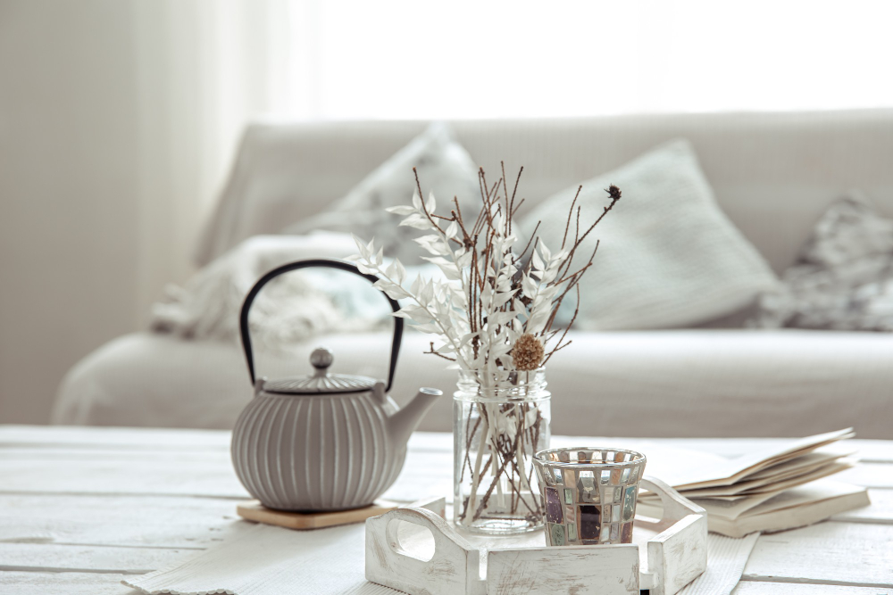 mesa baja blanca en sala con una pava blanca de metal y textura con una bandeja de madera pintada en blanco gastado, un vaso y un envase de vidrio con ramas secas decorativas.
