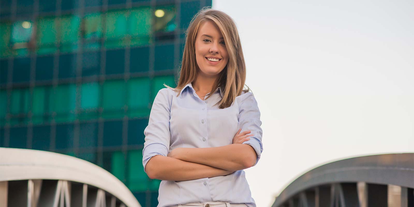 Mujer joven de brazos cruzados sonriendo, segura de sí misma, en un puente. Un puente que puede cruzar de un lado o del otro con el asesoramiento inmobiliario correcto.
