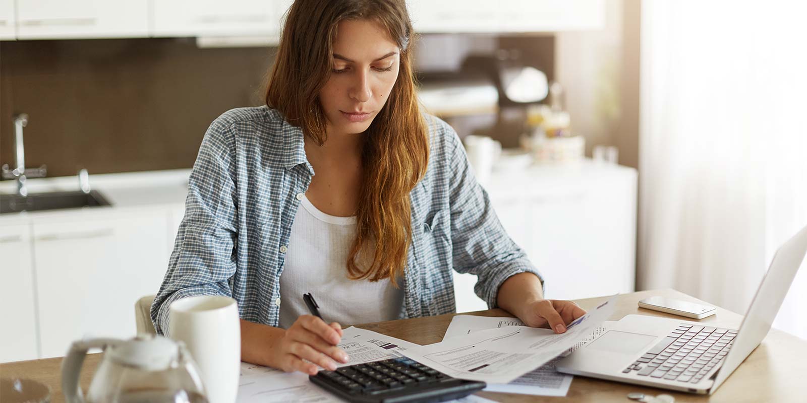 Mujer joven sentada en una mesa (dentro de una cocina) rodeada de documentación, una calculadora, un portátil con un bolígrafo en la mano y una jarra y una taza de café. En concepto de saber cuál es la ganancia patrimonial en el IRPF.