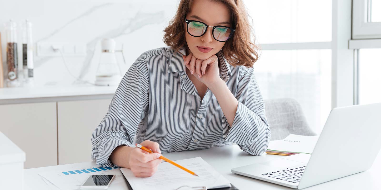 Mujer joven con gafas con lápiz en mano leyendo documentación y un portátil. En concepto de entender los tipos impositivos de Hacienda de la vivienda que podría vender.
