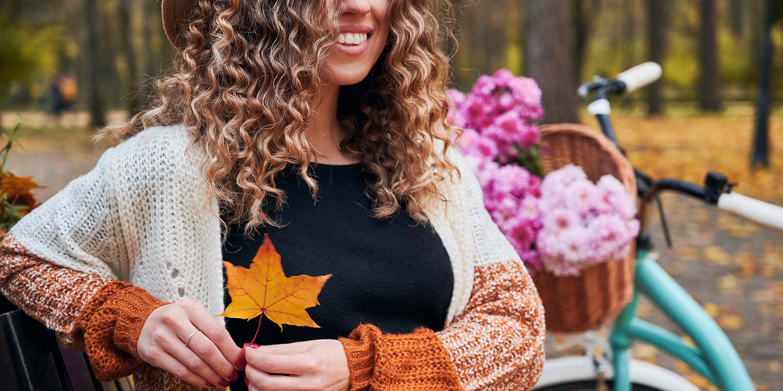 Imagen de mujer joven sonriendo sentada en un banco de un parque sosteniendo una hoja seca marrón de árbol y una bicicleta detrás de ella con una cesta con flores lilas. En representación de las estaciones Primavera y Otoño como las estaciones con más oportunidades para realizar operaciones inmobiliarias.