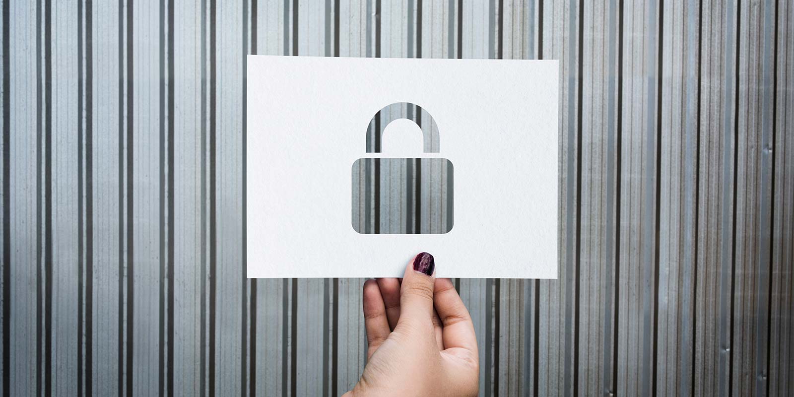 Imagen de mano de mujer sosteniendo un folio con una forma de candado recortado y de fondo una pared metálica. En referencia a la protección de la inversión inmobiliaria que realizas y las estrategias para reducir riesgos.