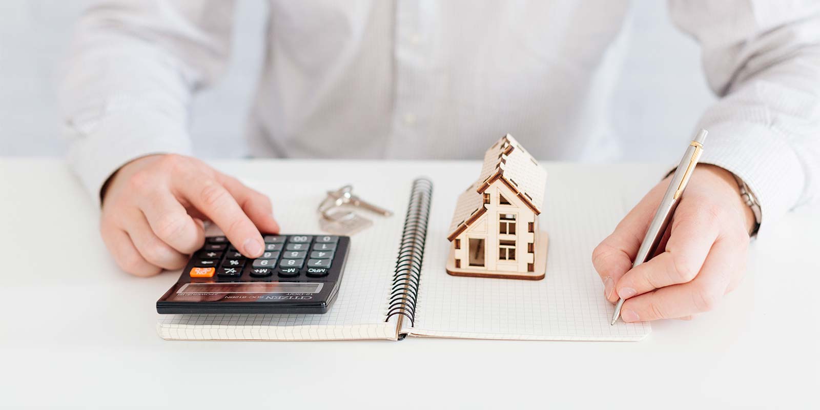 Imagen de zona media de hombre haciendo cuentas con una calculadora y el modelo de una casa sobre un escritorio. En referencia de qué hay que tener en cuenta a la hora de hacer la declaración de impuestos por el inmueble en la Costa del Sol,
