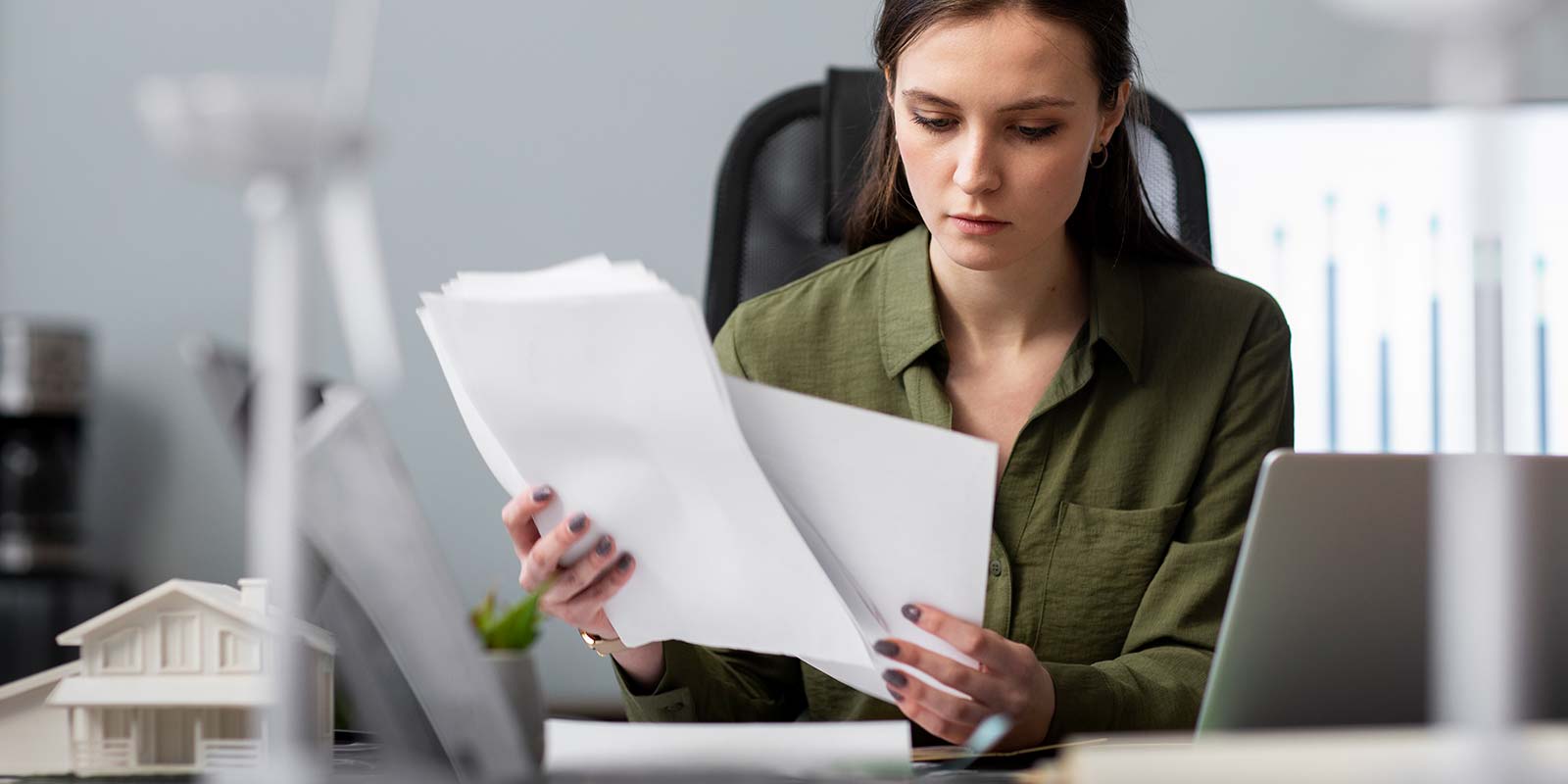 Mujer joven revisando documentación sobre escritorio con un modelo de casa sobre el escritorio. En referencia a la transparencia y confianza que un agente y asesor inmobiliario puede darle al cliente ante una oferta inmobiliaria en la Costa del Sol.